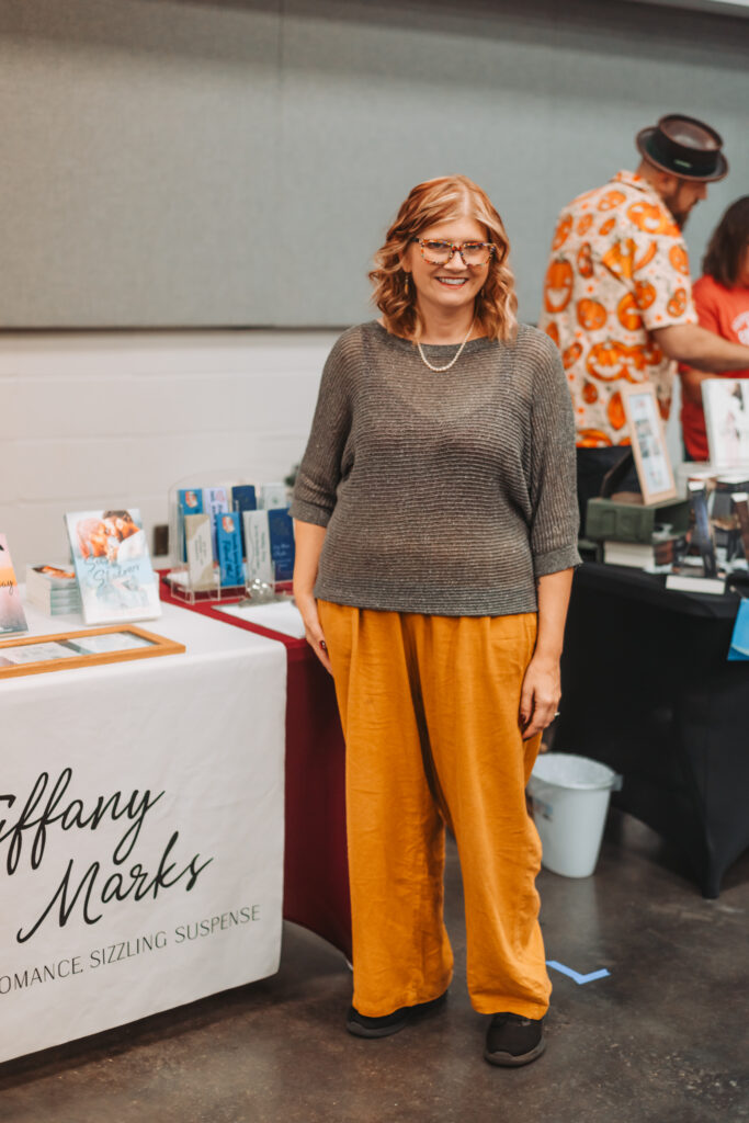 Tiffany Marks, contemporary romance author, smiles beside her Romance by TM display table at Foothills Book Fest in Jasper, Alabama.